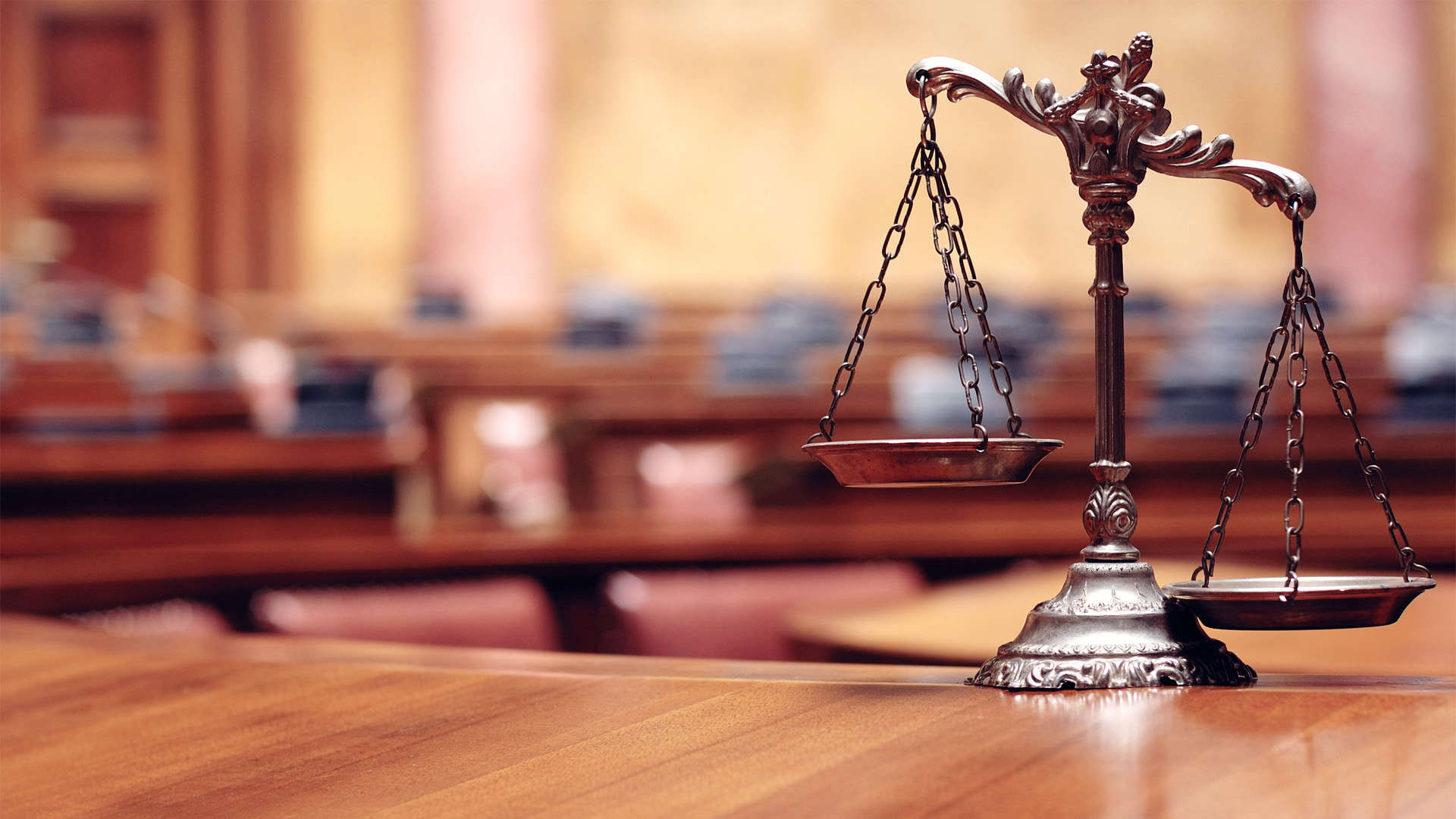 Scales of Justice on a table where the right is heavier than the left. The scene is in a courtroom where many rows of seating can be found but the background blurs.