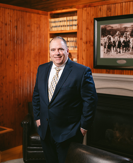 Tim Hayes of Hayes Law poses for a portrait inside of his office next to the fireplace and chairs. He is smiling proudlin in a nice suit and tie with hands placed in his pockets.